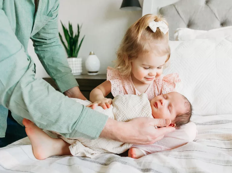 A toddler girl holds her newborn brother with daddy's help during newborn photos.