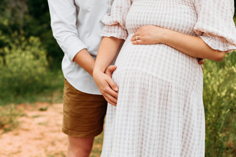 man in shorts with pregnant mom in pink check dress with baby bump during photo session at Arcadia Lake, Edmond, OK.