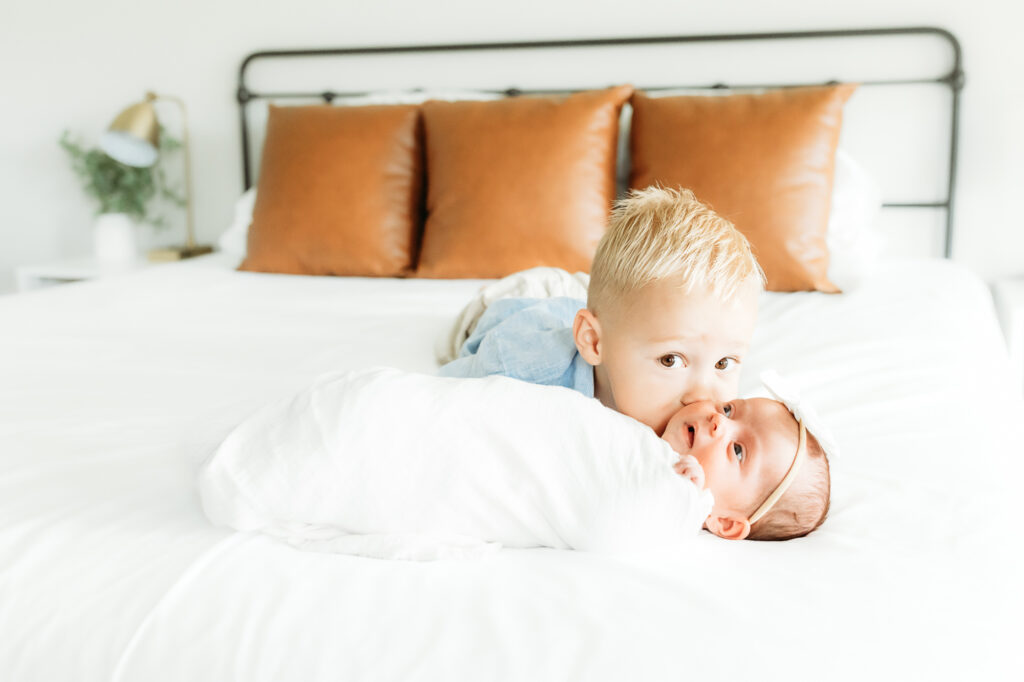 Toddler brother with newborn sister on bed with white comforter.