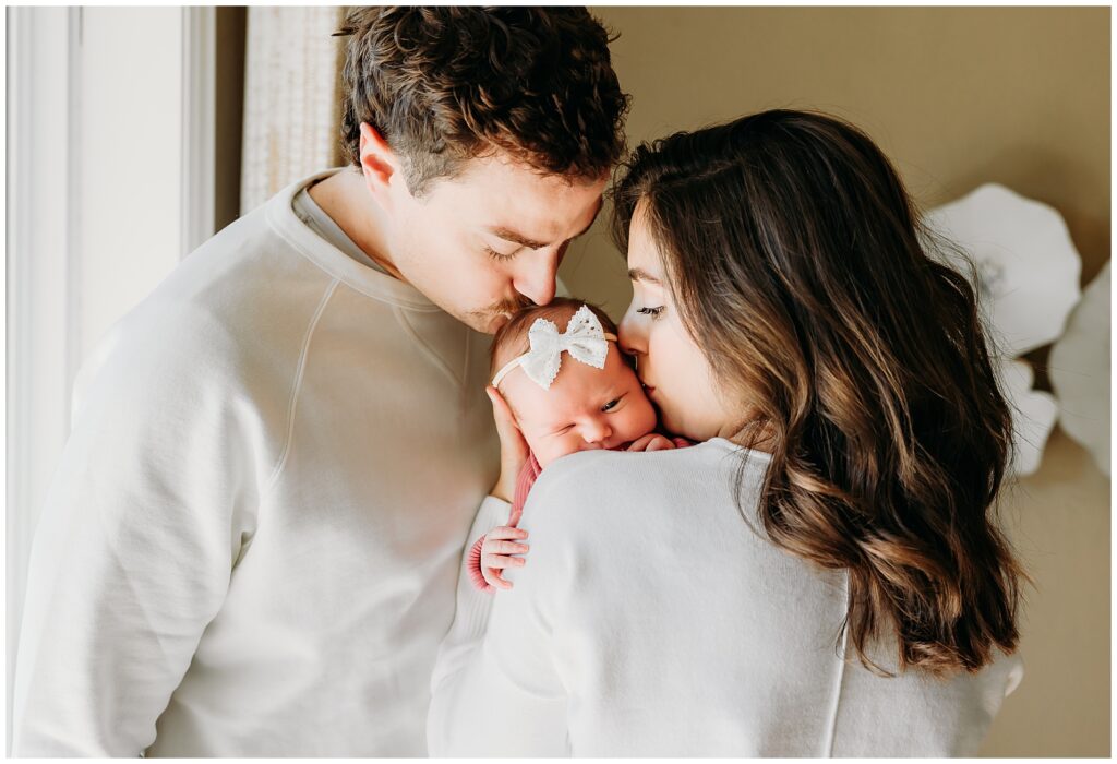 Mom and Dad kiss newborn daughter during in home newborn photo session.