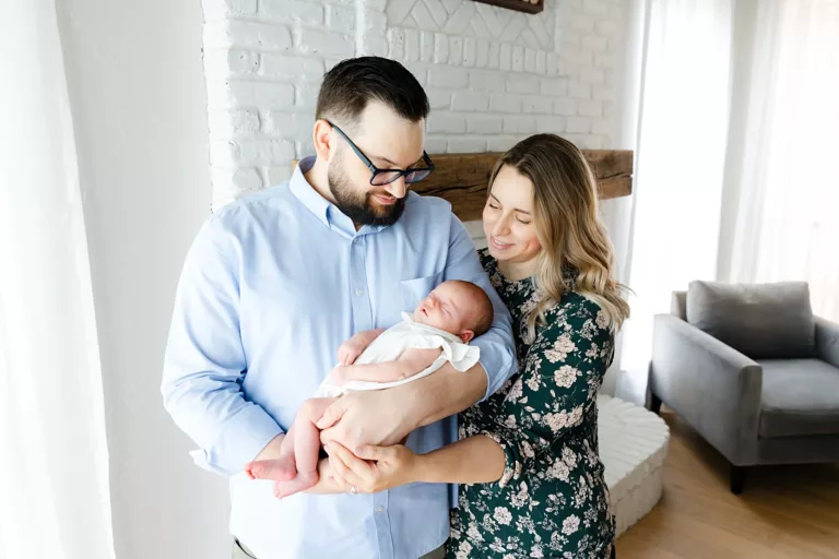 Dad in blue shirt and mom in green floral dress with baby in white dress during at home newborn photos.