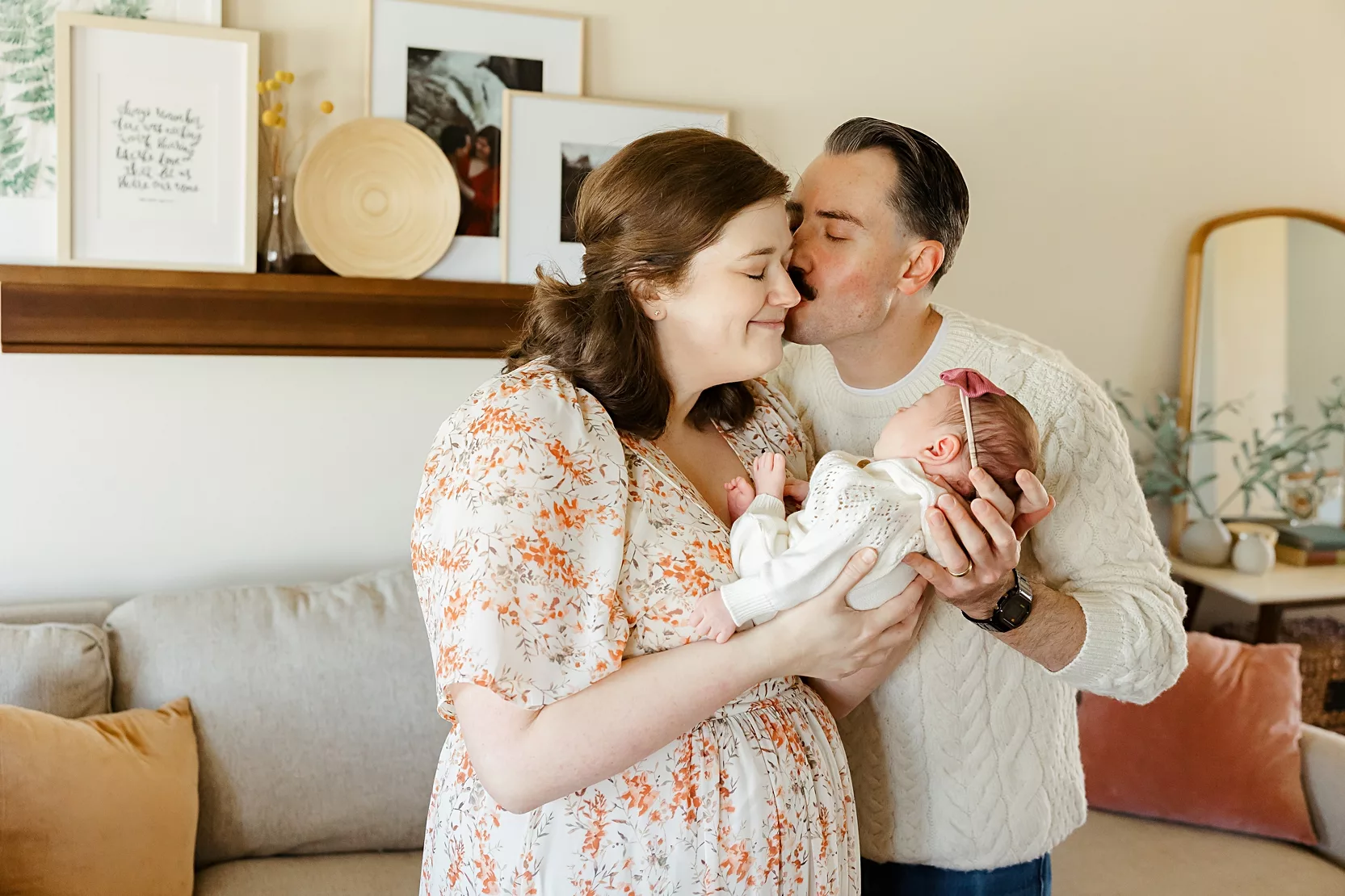 new parents stand in living room with mom in floral dress during newborn photos.